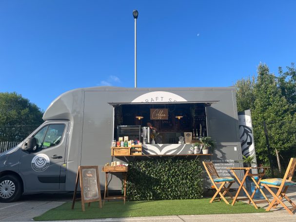 A grey food truck with a serving window, outdoor seating, and greenery nearby.