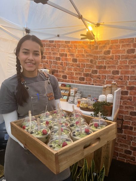 Smiling vendor holding a wooden tray with fruit-topped overnight oat pots ready for on-set smiles.