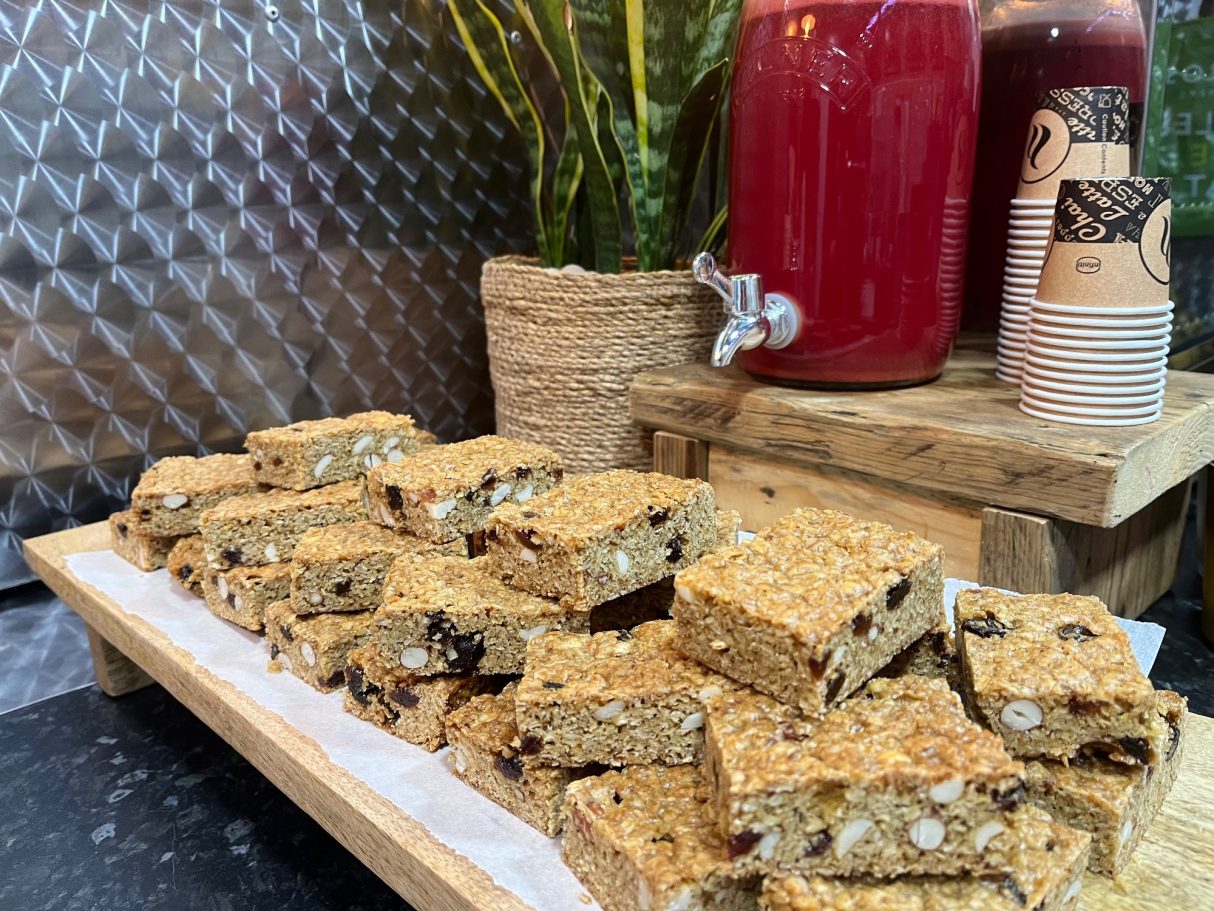 A wooden platter with sliced granola bars, decorated with jars of red beverages in the background.