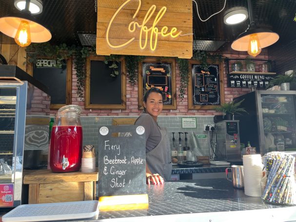 Barista at a coffee stand, with a menu board and decorative signage in the background.