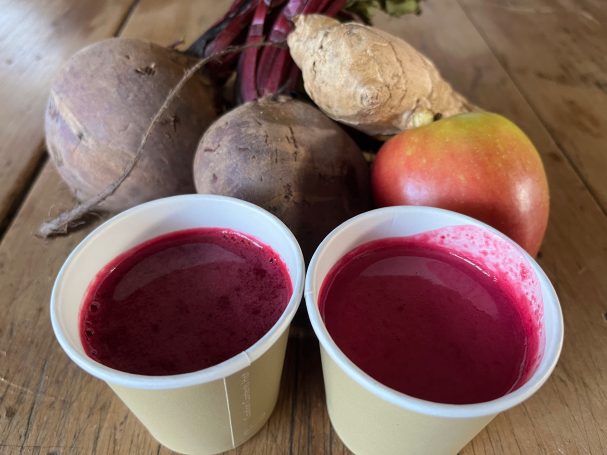 Two cups of vibrant red juice next to fresh root vegetables and an apple.