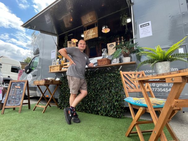 A vendor stands by a food truck with outdoor seating and greenery around.